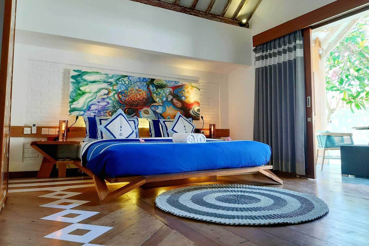 A modern bedroom with a large bed, a colorful abstract painting on the wall, and a view of the outdoors.