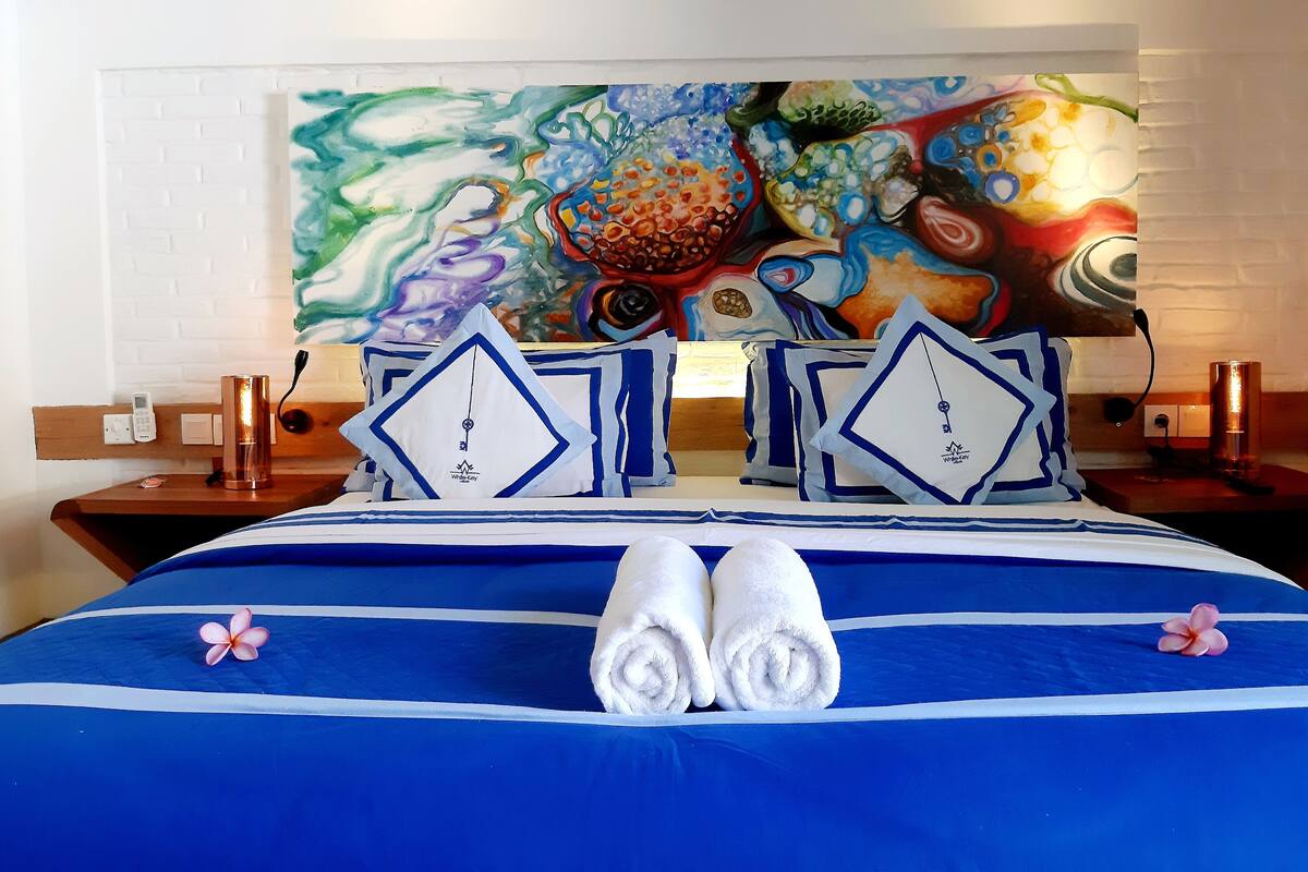 A hotel room with a bed featuring blue and white bedding, two pillows with a geometric design, and a colorful abstract painting on the wall behind the bed.