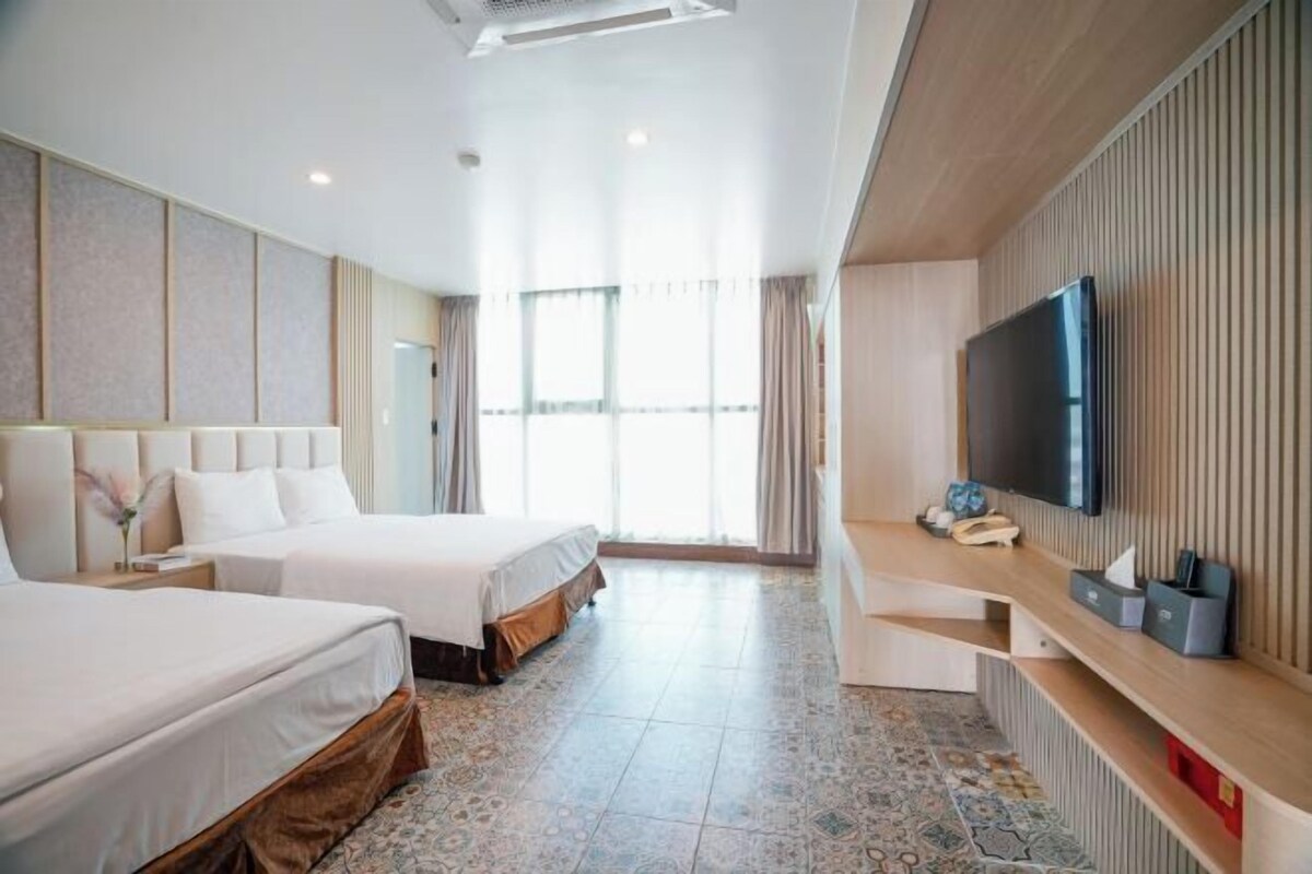 A hotel room with two beds, a TV, a desk, and a large window.
