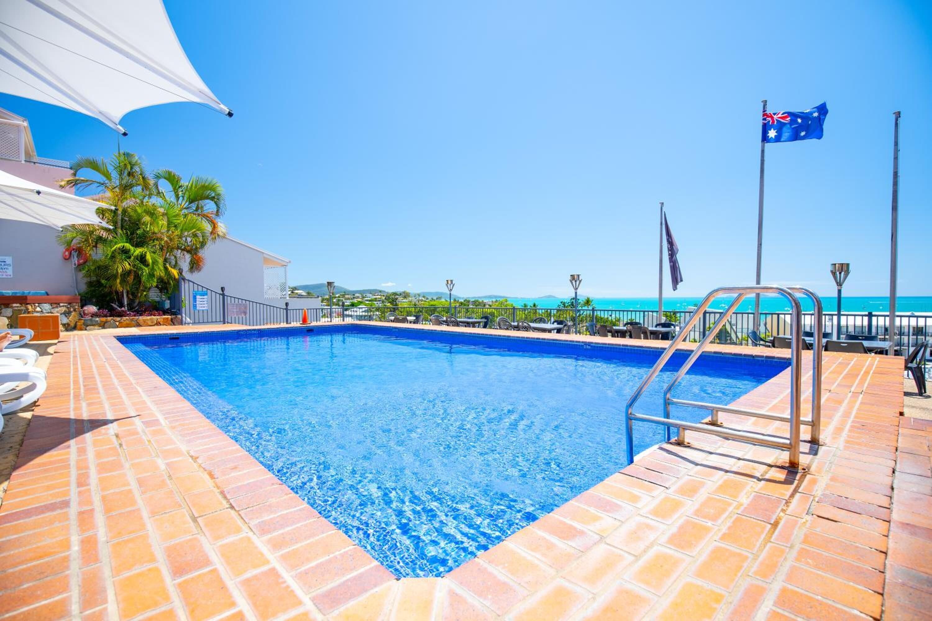 A swimming pool with a clear blue water, surrounded by a tiled deck, and a flagpole with a flag.