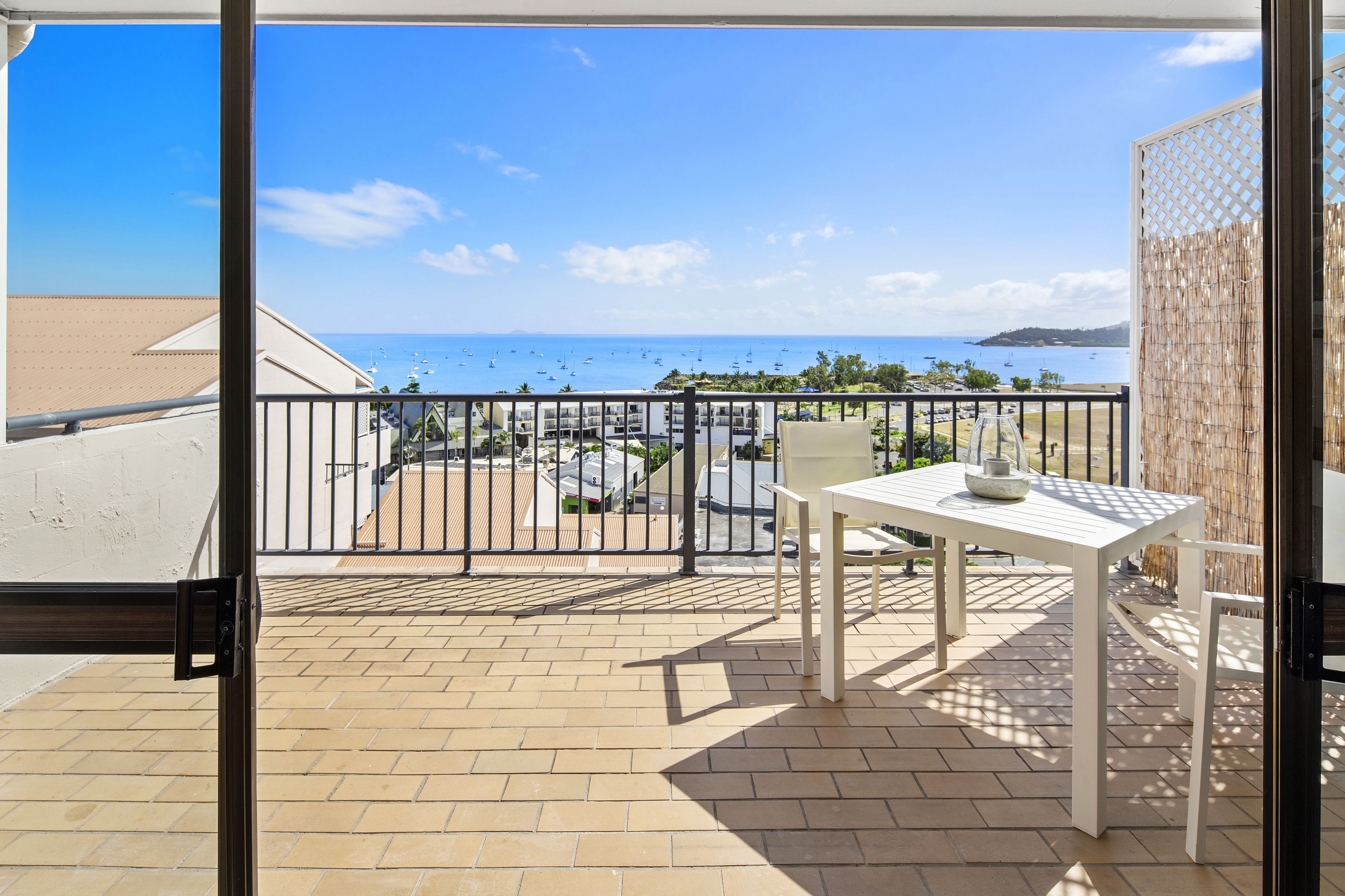 Uma varanda com mesa e cadeiras brancas, vista para o mar e uma área de estacionamento ao fundo.