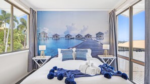 Quarto de hotel com uma cama grande, vista para bangalôs sobre a água e uma varanda com vista para palmeiras.