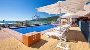 Piscina com espreguiçadeiras e guarda-sóis, área de bar e vista para o mar.