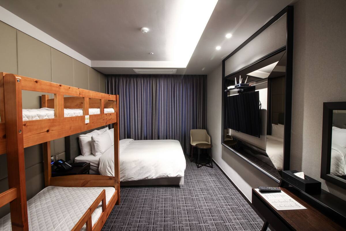 A hotel room with bunk beds, a single bed, a desk with a computer, a chair, a mirror, and a window with curtains.