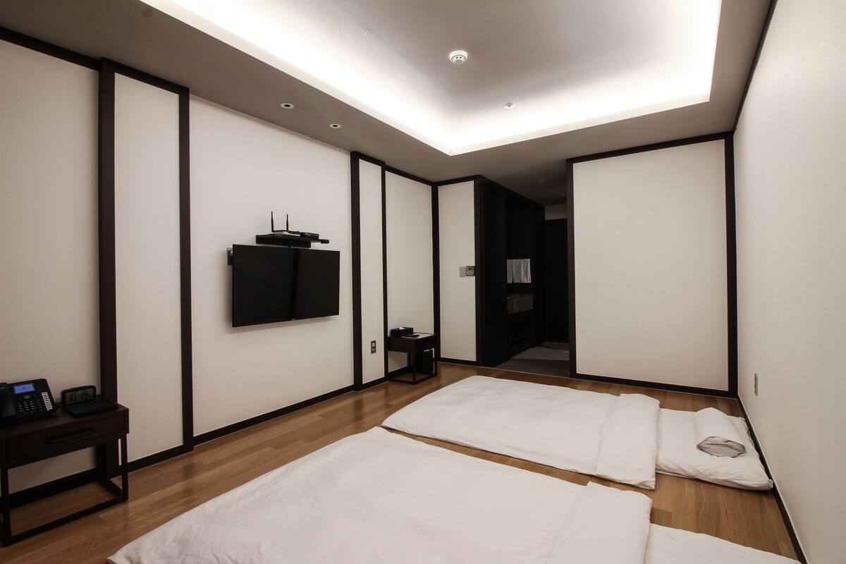 A modern hotel room with a wooden floor, two beds, a flat-screen TV mounted on the wall, and a small bedside table with a phone and a lamp.
