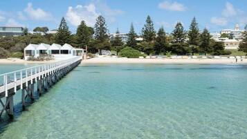 A clear waterfront with a wooden pier leading to a beach, surrounded by trees and buildings.