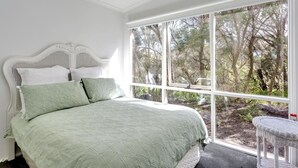 A bedroom with a large window, a white bed with green bedding, and a wicker chair. - The Heyfield Hideaway Rye (RYE)