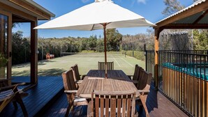 An outdoor deck with a wooden table and chairs, a white umbrella, and a tennis court in the background. - The Heyfield Hideaway Rye (RYE)