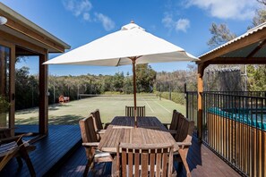 A wooden deck with a table and chairs, a white umbrella, and a tennis court in the background. - The Heyfield Hideaway Rye - PAY 2, STAY 3 Nights! (RYE)