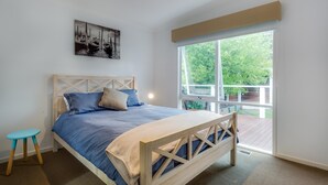 Un dormitorio con cama, un taburete pequeño, una ventana con vista a follaje y un cuadro en la pared.