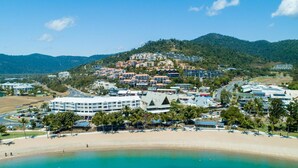 Un resort costero con villas frente a la playa, un hotel central y montañas al fondo.