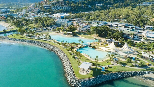 Vista aérea de un complejo turístico costero con varias piscinas, un litoral rocoso y edificios residenciales al fondo.