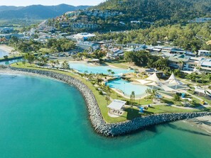 An aerial view of a coastal resort with multiple swimming pools, a rocky shoreline, and a backdrop of residential buildings. - Ocean Views 14 (Airlie Beach)