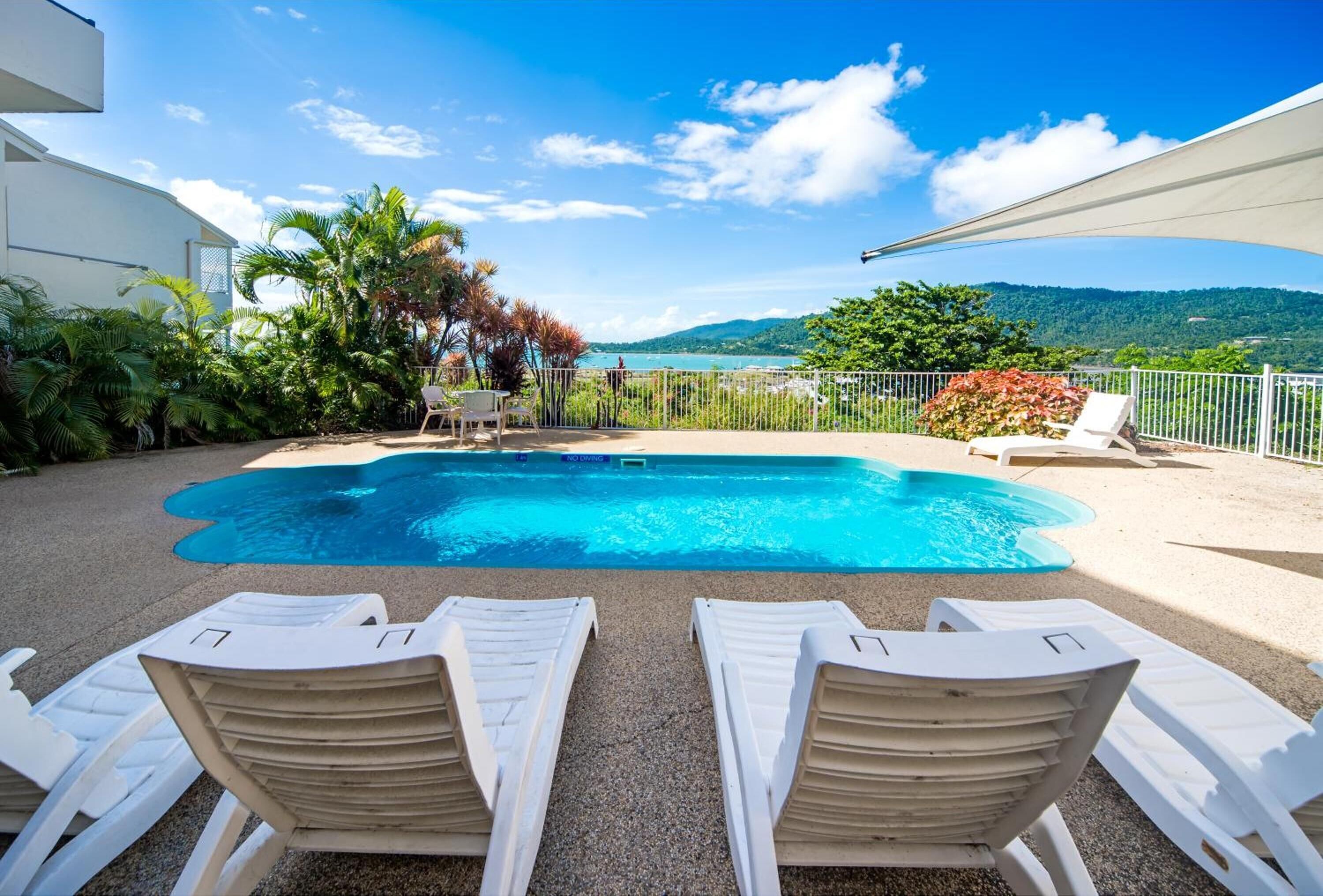 A swimming pool with white lounge chairs, surrounded by greenery and a view of a lake and mountains.