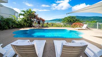 A swimming pool with white lounge chairs, surrounded by greenery and a view of a lake and mountains.