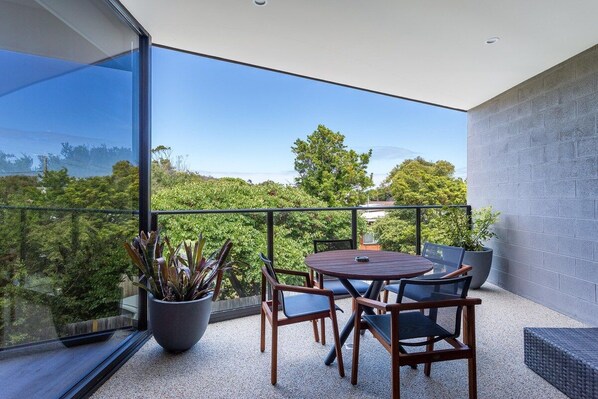 Ein moderner Balkon mit Glasveranda, einem runden Holztisch und Stühlen, umgeben von Grünpflanzen und freiem Blick auf die Landschaft.