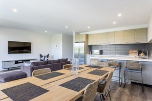 A modern kitchen with built-in appliances, a dining area with a wooden table and wicker chairs, and a living area with a sofa and a flat-screen TV. - Blairgowrie Apartment 2 (Blairgowrie)