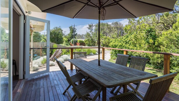 An outdoor deck with a wooden table and chairs, a large umbrella, and a view of a garden.