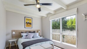 A bedroom with a large window, a ceiling fan, a bed with a headboard, and a framed poster on the wall.