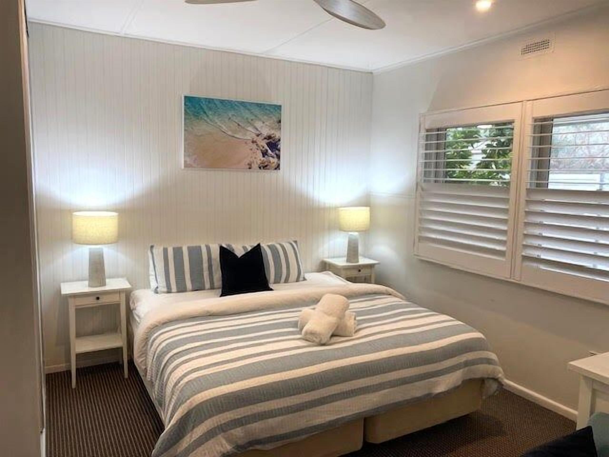 A hotel room with a bed, two bedside lamps, a window with blinds, and a wall-mounted artwork.