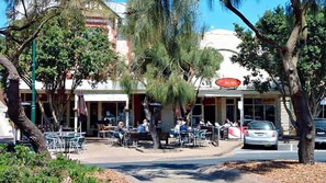 Un café al aire libre con mesas y sillas, árboles, y un edificio con toldo rojo al fondo.
