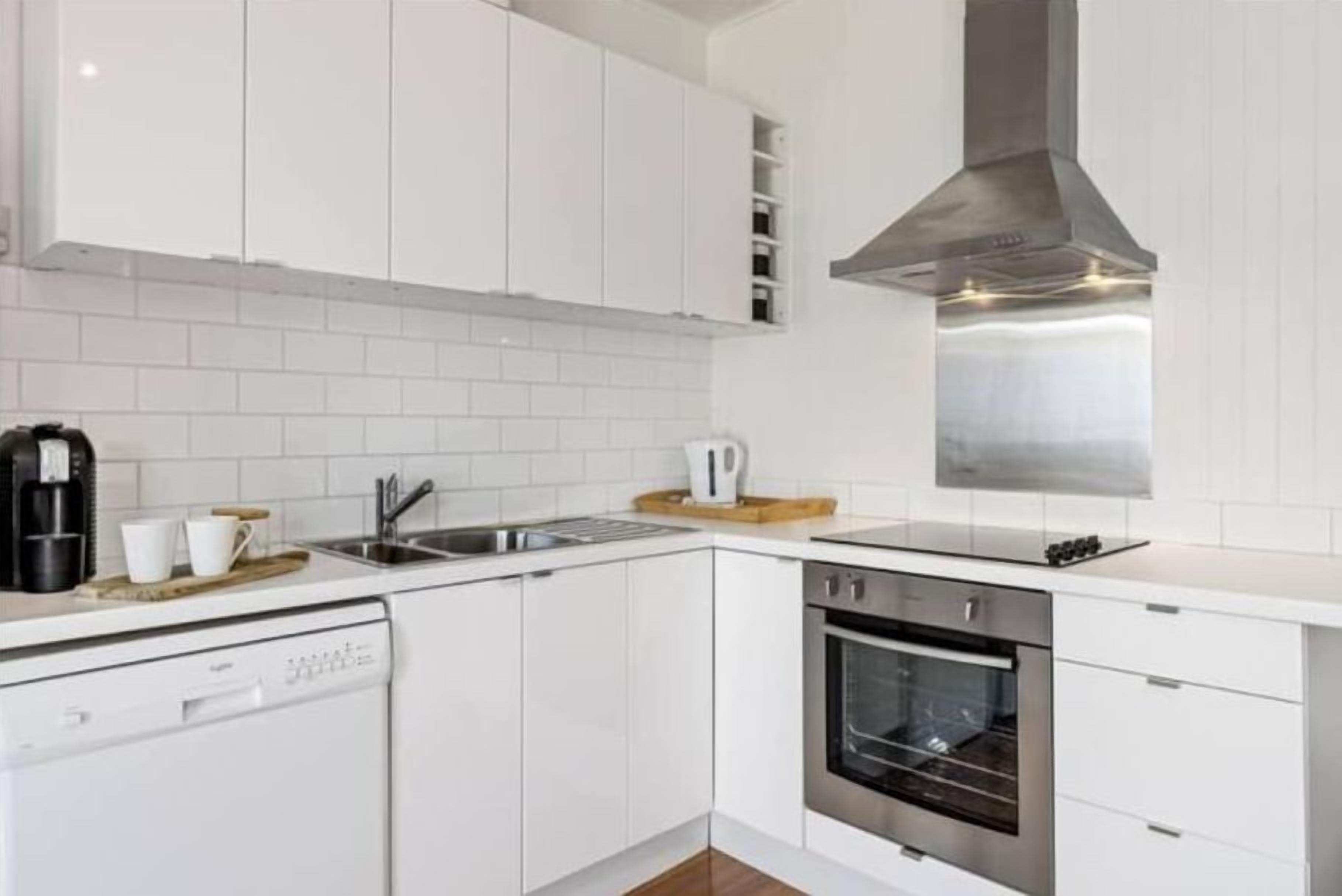 A modern kitchen with white cabinetry, a stainless steel range hood, a dishwasher, and an oven.