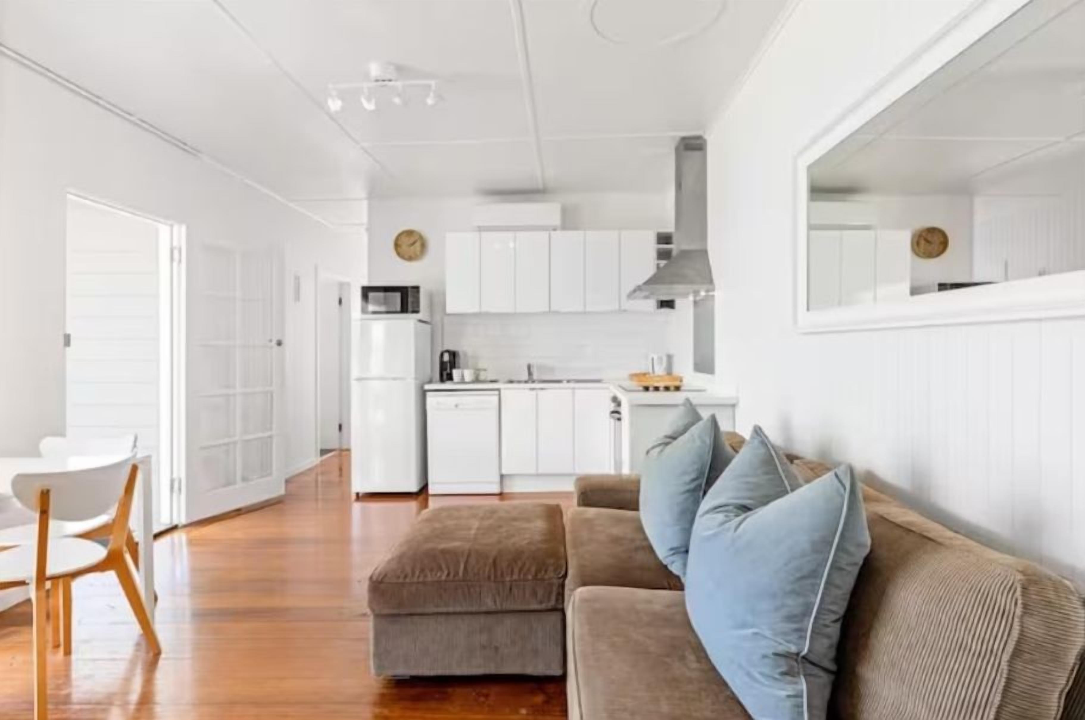 A modern kitchen with white cabinets, a microwave, and a stove. There is a dining area with a white table and chairs. A brown sofa with blue cushions is against the wall.
