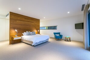 Een moderne slaapkamer met een groot bed, nachtkastjes, een blauwe fauteuil, een klein krukje en een televisie aan de muur.