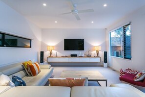 Een moderne woonkamer met een flatscreen televisie, twee bijzettafels met lampen, een plafondventilator en een groot raam.