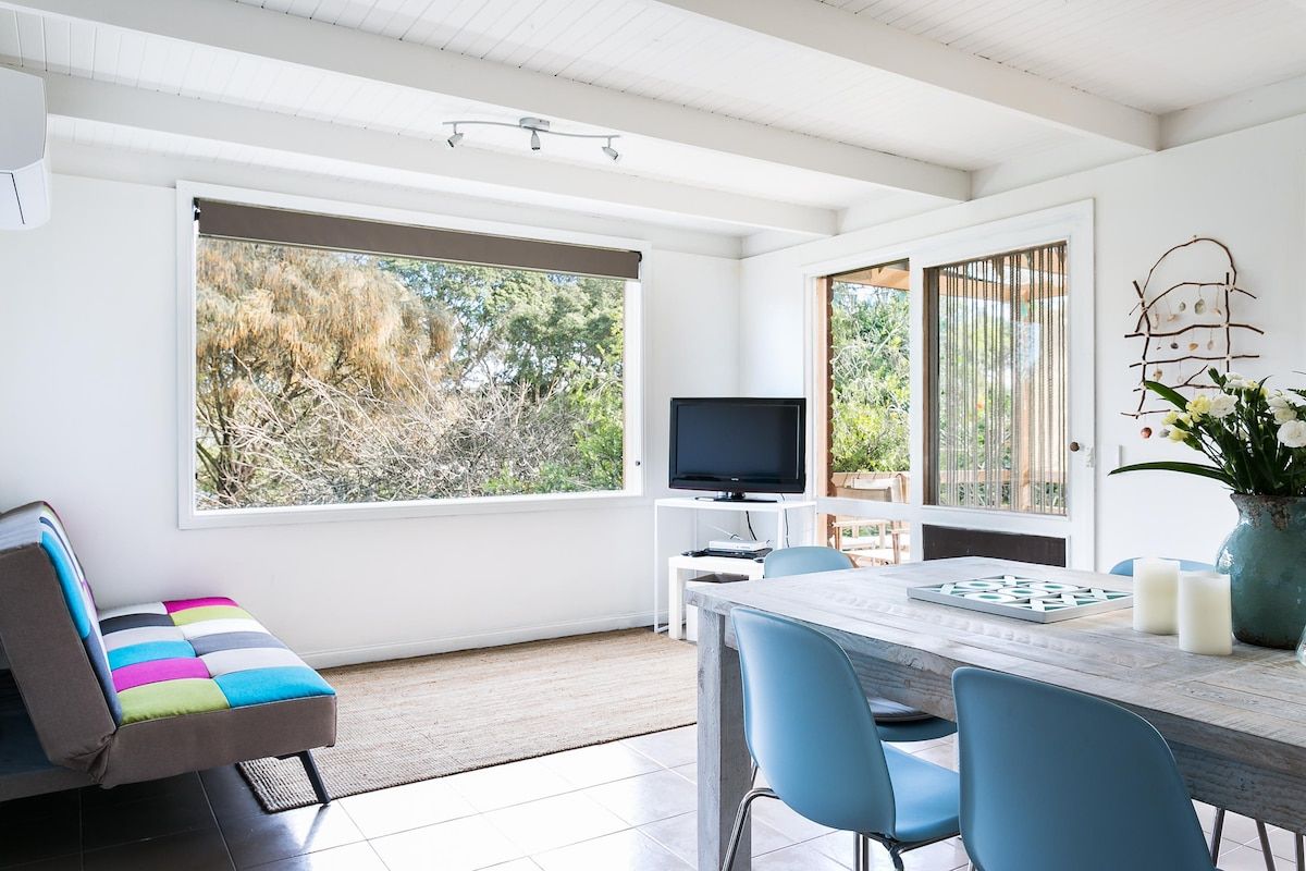 Un salon moderne doté d’une grande fenêtre, d’un téléviseur à écran plat, d’une table à manger avec des chaises et d’un canapé aux couleurs vives.