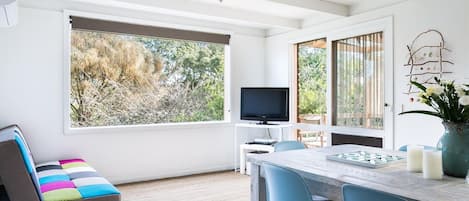Een moderne woonkamer met een groot raam, een flatscreen televisie, een eettafel met stoelen en een kleurrijke bank.