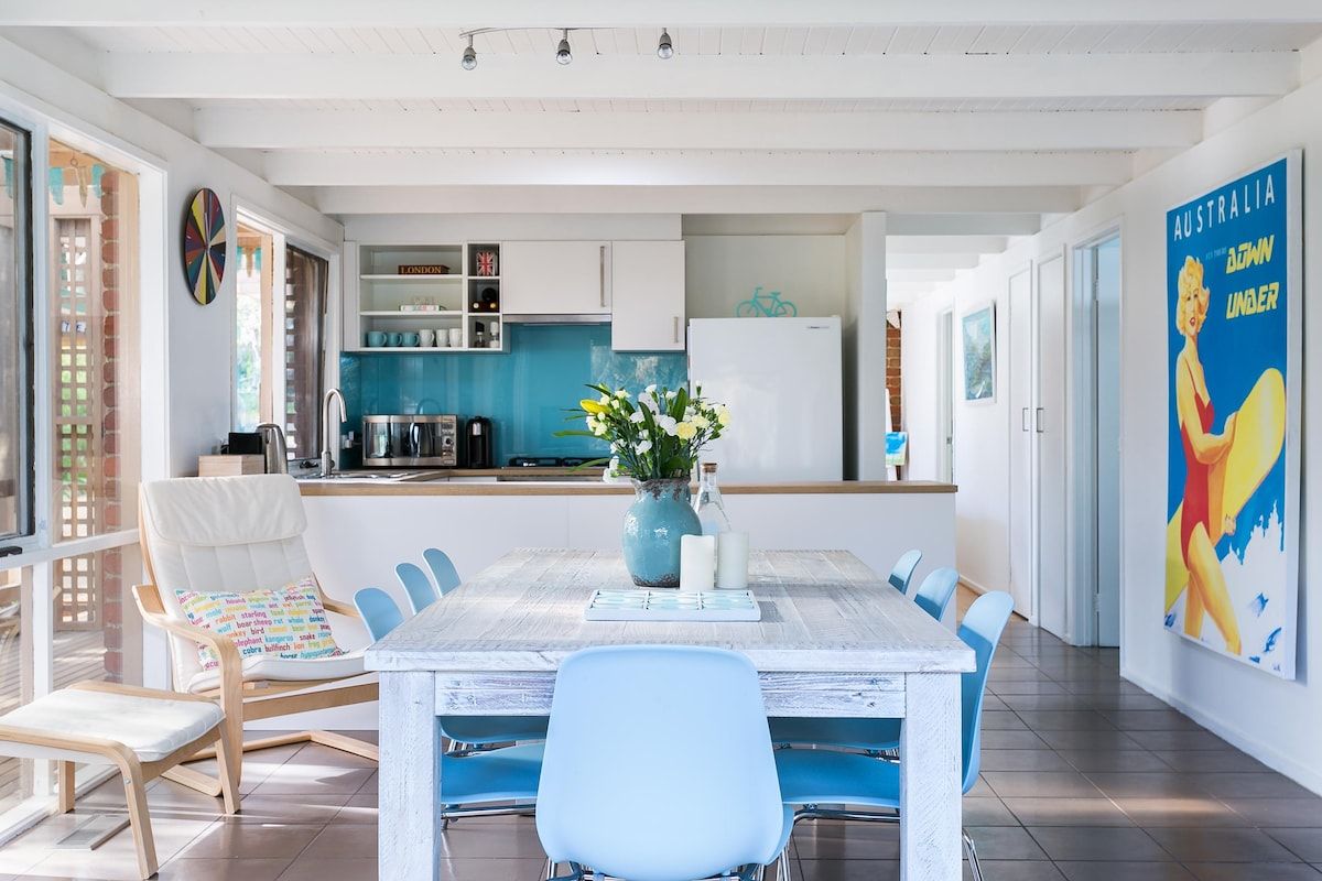 Een moderne keuken met witte kasten, een blauwe achterwand en een eethoek met een witte tafel en blauwe stoelen.
