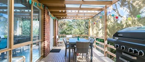 An outdoor patio with a wooden deck, a table with chairs, a grill, and a glass enclosure with a view.