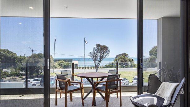 A balcony with a table and chairs, offering a view of the sea and a parking area.