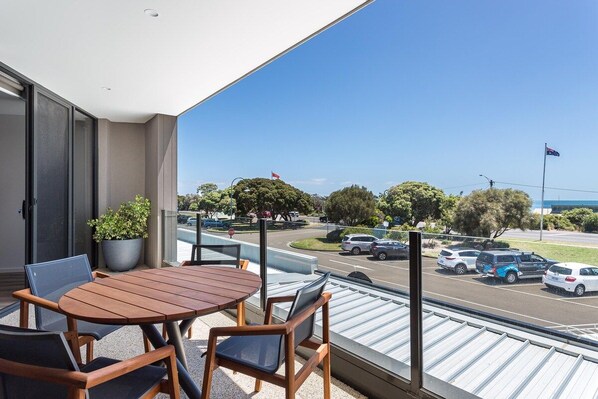 A balcony with a wooden table and chairs, a potted plant, and a view of a street with cars and trees. - Blairgowrie Apartment 1 (Blairgowrie)