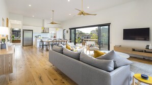 A modern living room with a grey sofa, wooden flooring, ceiling fans, a dining area with chairs, and a TV on the wall. - Yaringa, St Andrews Beach - Pristine new family living and only moments from the surf beach (ST ANDREWS BEACH)