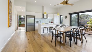 A modern kitchen with white cabinets, a central island with stools, and a dining area with a large wooden table and chairs. - Yaringa, St Andrews Beach - Pristine new family living and only moments from the surf beach (ST ANDREWS BEACH)