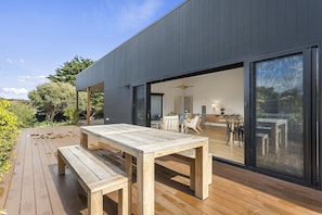 A modern house with a wooden deck, a dining area with chairs and a table, and a large sliding glass door leading to an interior space.