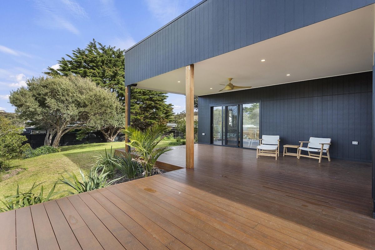 Une terrasse extérieure moderne avec un revêtement de sol en bois, un espace salon couvert avec des chaises, et une vue sur un jardin aménagé avec des arbres et des arbustes.