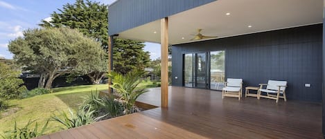 A modern outdoor deck with wooden flooring, a covered seating area with chairs, and a view of a landscaped garden with trees and shrubs.