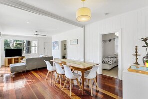 A spacious living area with a wooden dining table, white chairs, a grey sofa, and a flat-screen TV. - Sorrento Beach House. (SORRENTO)