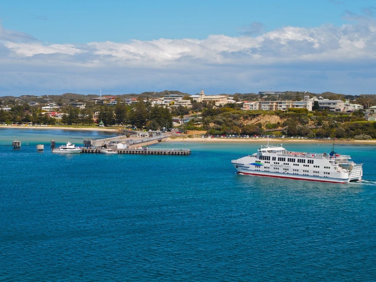 A ferry boat with multiple decks and a prominent name on its side, docked at a pier with a clear view of a coastal town and hills in the background.
