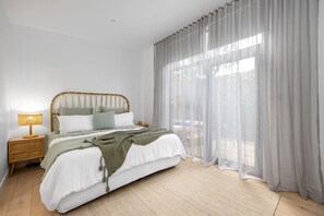 A bedroom with a bed, a nightstand, a lamp, sheer curtains, and a view of the outdoors.