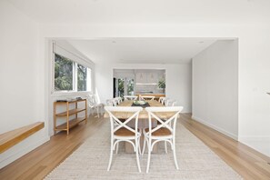 A spacious dining area with a wooden table and white chairs, a kitchen in the background, and a large window providing natural light.