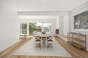 Un comedor moderno con una mesa de madera y sillas blancas, una chimenea y vistas a palmeras a través de amplios ventanales.