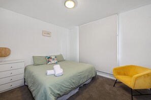 Une chambre à coucher avec un lit, une chaise jaune, une commode et un luminaire fixé au mur.