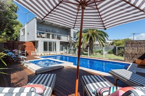 Une piscine entourée d’une terrasse en bois et d’un bâtiment à deux étages, avec des chaises longues et un parasol à proximité.