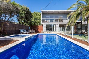 A swimming pool with a wooden deck, a covered patio area, and a modern two-story house with large windows.