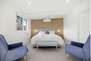 A bedroom with a bed, two blue chairs, a ceiling fan, and a window with a view.
