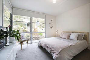 A bedroom with a bed, a chair, a desk with a plant, and a window with a view of the outside.